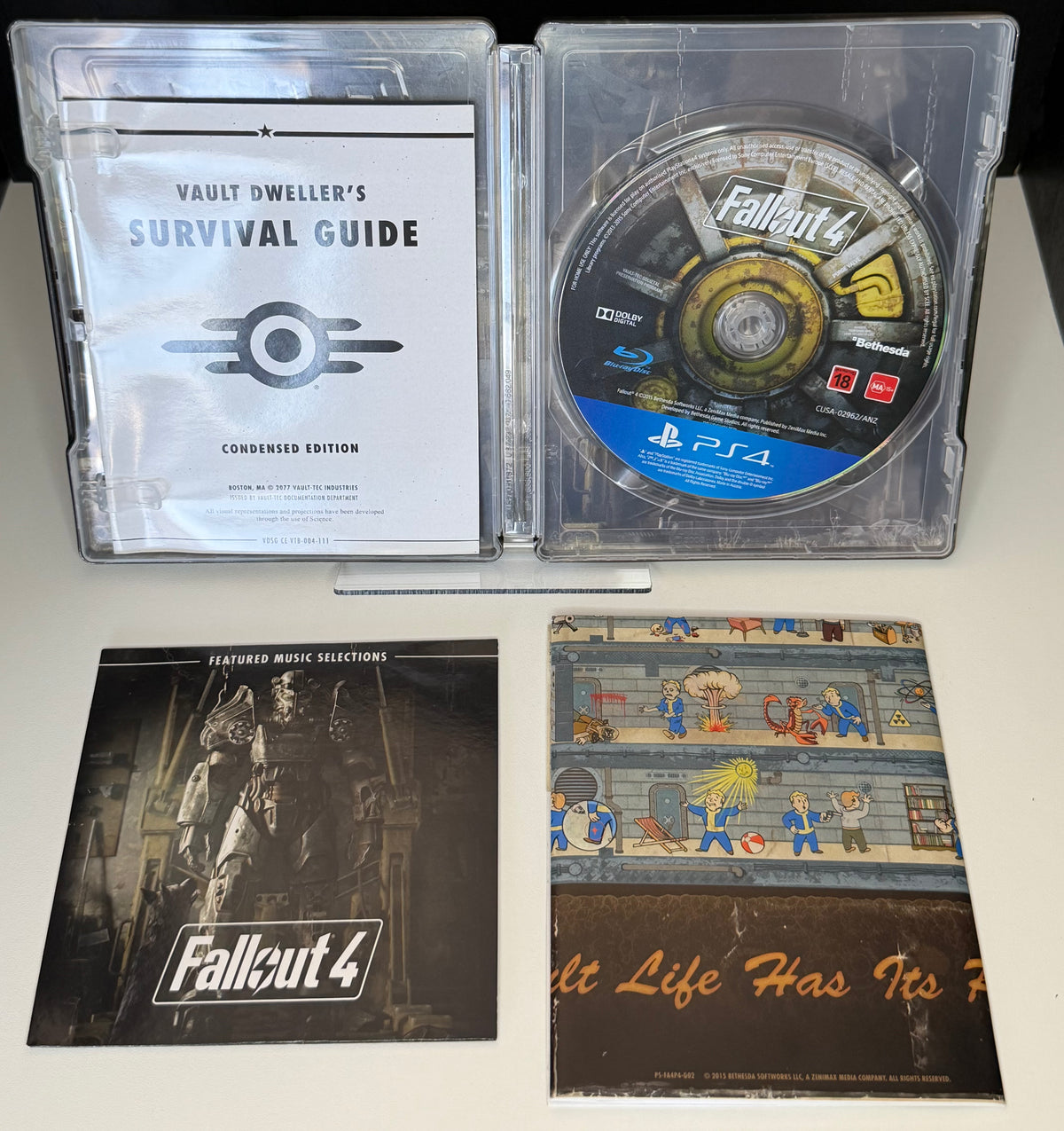 Fallout 4 SteelBook Edition PS4 open case showing Vault Dwellers Survival Guide Condensed Edition inside SteelBook with game disc and music card and Vault Boy booklet laid out below