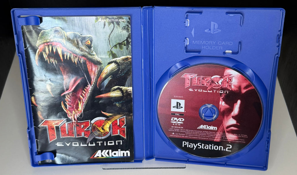 Turok Evolution PS2 open case showing AML 23273.01 ENG manual with T-Rex artwork on left panel and PAL DVD-ROM Acclaim game disc in right tray