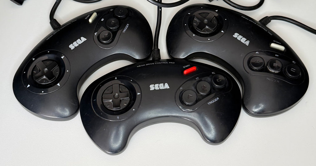 Four official Sega Mega Drive Control Pad 3-button wired controllers in black showing varying cosmetic wear consistent with Grade B pre-owned condition