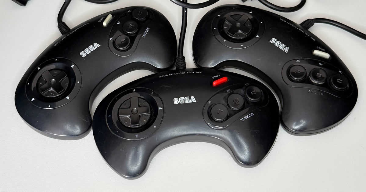 Four official Sega Mega Drive Control Pad 3-button wired controllers in black showing varying cosmetic wear consistent with Grade B pre-owned condition
